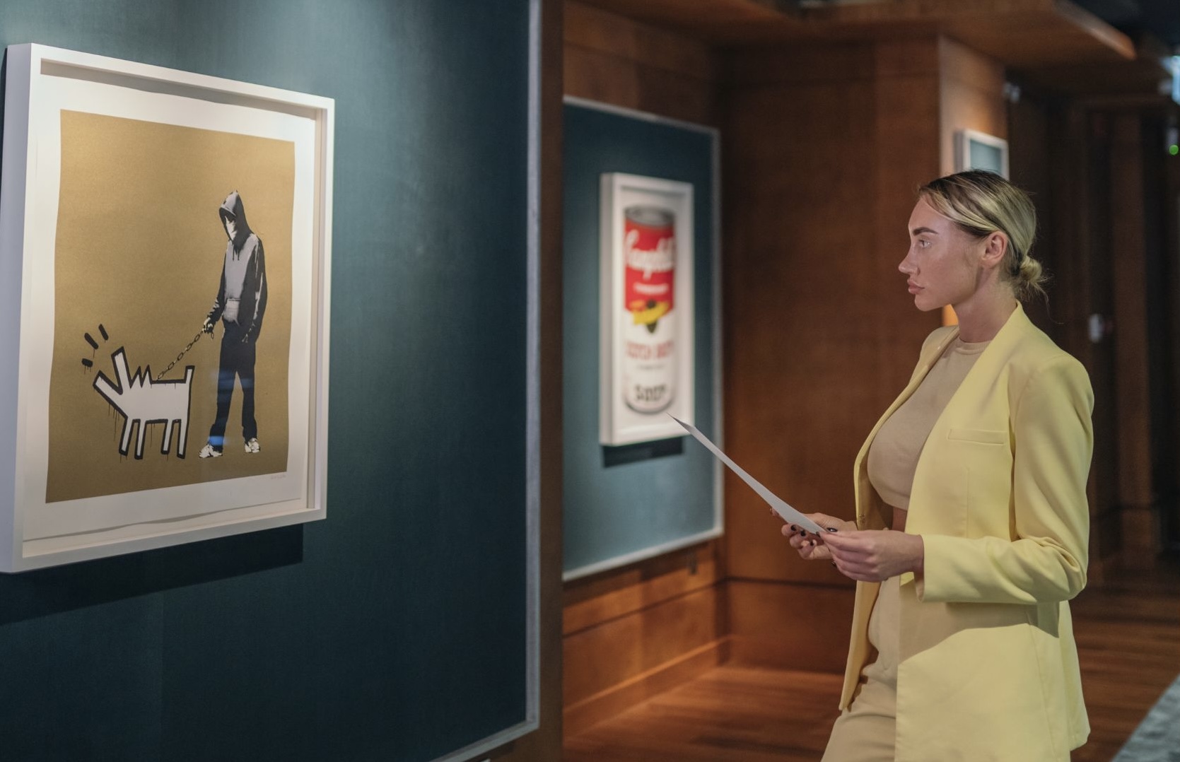 A woman attentively reads a document while standing in an art gallery with dark walls. Behind her, two iconic framed artworks are displayed: a Banksy painting depicting a hooded figure walking a geometrically abstract dog on a gold background, and Andy Warhol's 'Campbell's Soup' can.